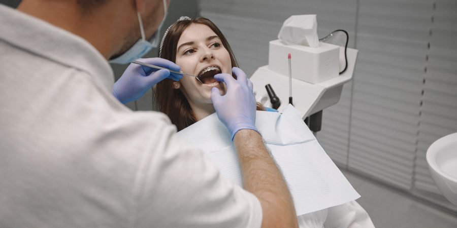 Female patient with braces has dental examination at dentist office. Woman wearing white clothes. Male dentist treats girl.